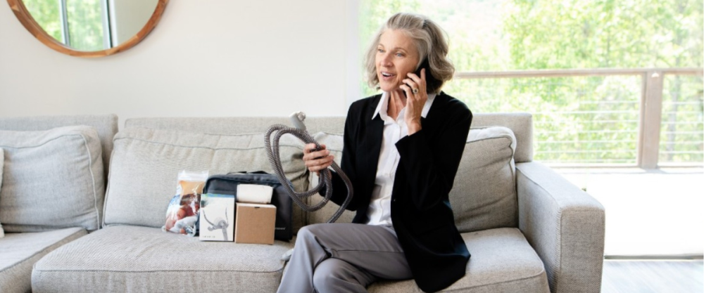 Woman holding CPAP machine tubing with other CPAP products around her and talking on the phone