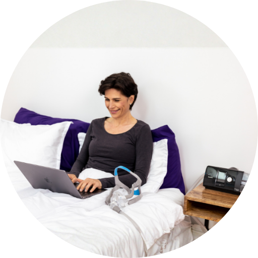 woman in bed on the computer with a full face cpap mask and a resmed cpap machine on her nightstand