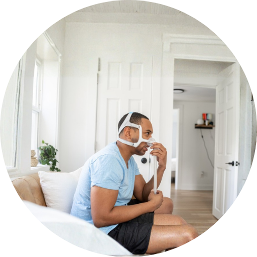 Man with facial hair sitting on a couch wearing a nasal CPAP mask used for CPAP therapy in sleep apnea patients